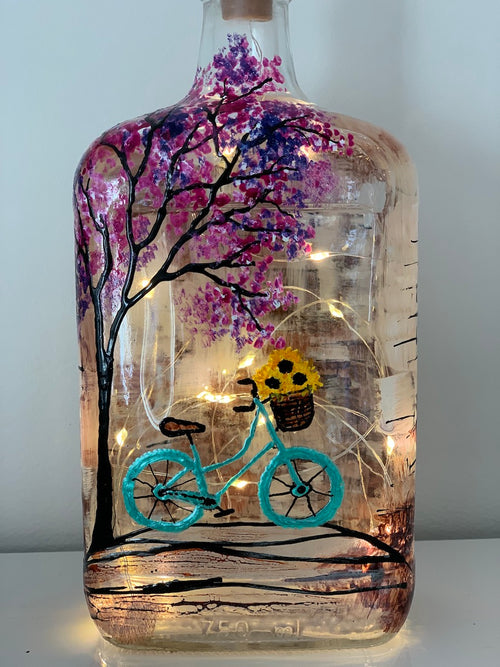 A blue bicycle with yellow flowers in the basket rides along the ground in front of an interesting wall and under a cherry blossom tree. Painted on a recycled bottle with acrylic paint. The bottle is lit with fairy lights from within.