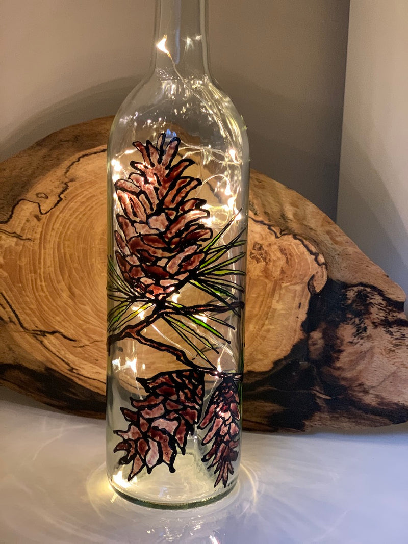 Open pinecones adorn a single pine branch that wraps around this bottle. Painted on a recycled bottle with acrylic paint. The bottle is lit with fairy lights from within.