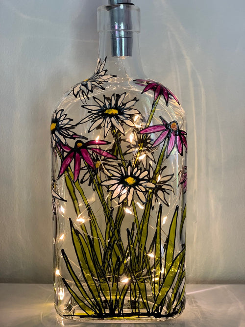 White and purple flowers with green stalks painted on a recycled bottle with acrylic paint. The bottle is lit with fairy lights from within.