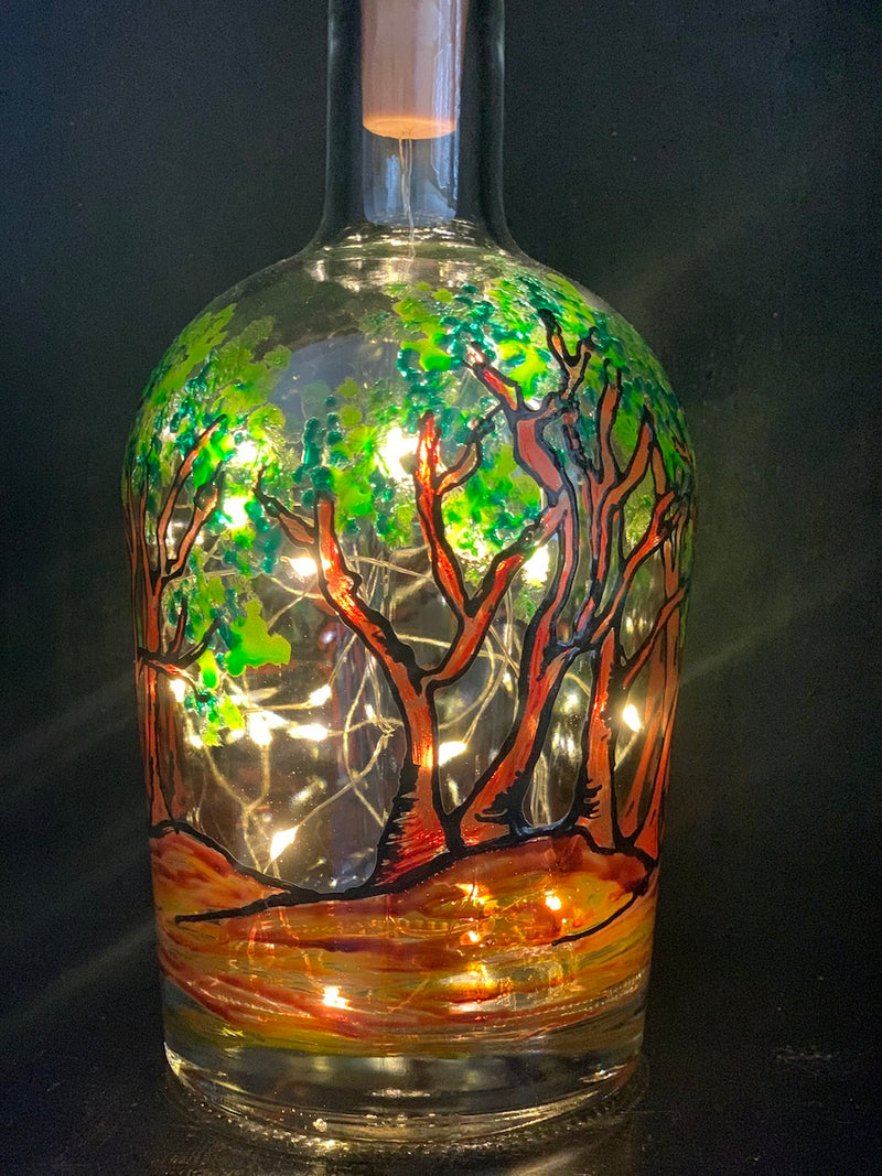 A grove of arbutus trees painted on a recycled bottle with acrylic paint. The bottle is lit with fairy lights from within.
