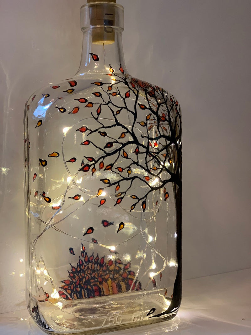 A tree in the corner of this flask shaped bottle is starting to drop leaves. Red, yellow and orange leaves fall from the branches and have formed a pile at the bottom of the bottle. Painted on a recycled bottle with acrylic paint. The bottle is lit with fairy lights from within.