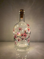 Pink blossoms on a branch that winds around a clear bottle. Painted on a recycled bottle with acrylic paint. The bottle is lit with fairy lights from within.