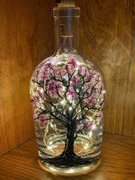 A dark tree in full bloom with blossoms on a clear whiskey bottle. Painted on a recycled bottle with acrylic paint. The bottle is lit with fairy lights from within.