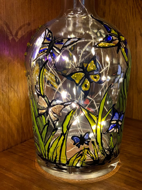 Many-coloured butterflies flying in a meadow. Painted on a recycled bottle with acrylic paint. The bottle is lit with fairy lights from within.