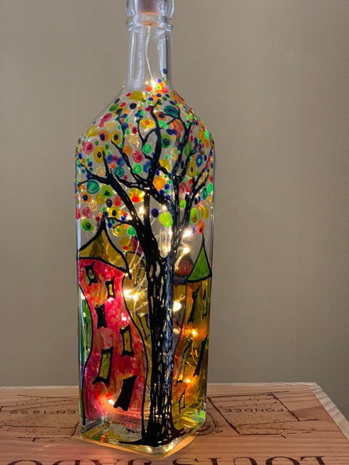A tree with multi-coloured button shaped leaves stands amidst a village of wavy-shaped colourful buildings. Painted on a recycled bottle with acrylic paint. The bottle is lit with fairy lights from within.