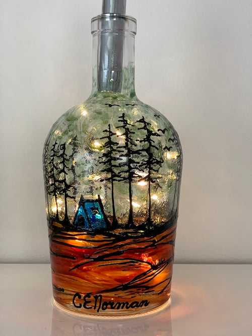 A blue tent sits amidst a grove of trees while birds fly above. Painted on a recycled bottle with acrylic paint. The bottle is lit with fairy lights from within.