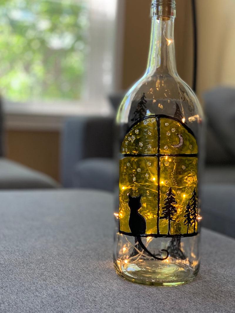 A black cat is sitting, silhouetted in front of a large window that glows yellow.  Trees and stars can be seen through the window. Painted on a recycled bottle with acrylic paint. The bottle is lit with fairy lights from within.