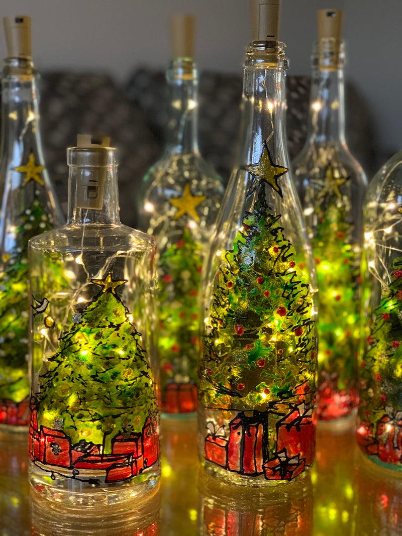 A decorated Christmas tree with large star on top is full of wrapped red presents underneath. Painted on a recycled bottle with acrylic paint. The bottle is lit with fairy lights from within.