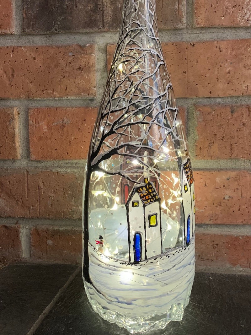 A winter scene has a small village with homes being warmed by their fireplace on a snowy night under large trees. A snowman seems to be running along the snow. Painted on a large magnum recycled bottle with acrylic paint. The bottle is lit with fairy lights from within.