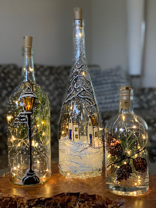 A trio of bottles includes the Christmas Village Magnum, Narnia Lantern, and Pinecones. Painted on a recycled bottle with acrylic paint. The bottles are lit with fairy lights from within.