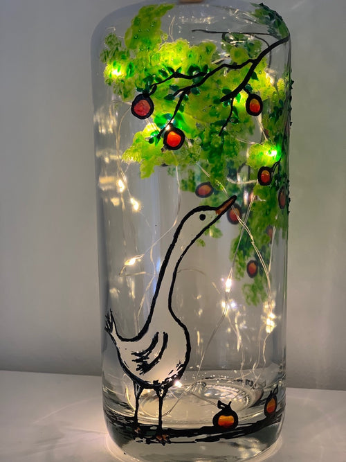 A large white hungry goose reaches to the apple tree for one of the many red apples found there. Some apples lie about on the ground. Painted on a recycled bottle with acrylic paint. The bottle is lit with fairy lights from within.