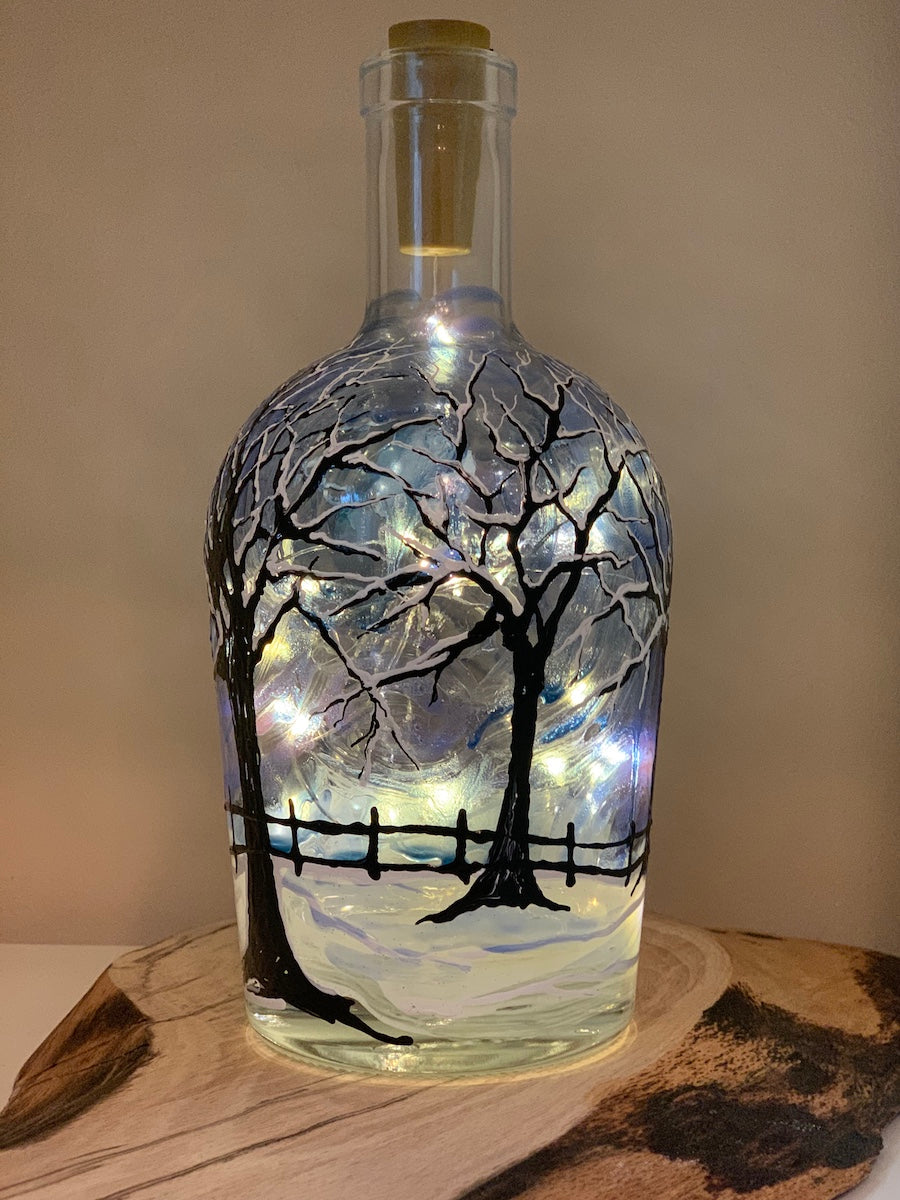 In the bleak midwinter is a snowy night with dark trees covered in snow, in a field with a fence. A full moon shines down through a sliver blue night sky. Painted on a recycled bottle with acrylic paint. The bottle is lit with fairy lights from within.