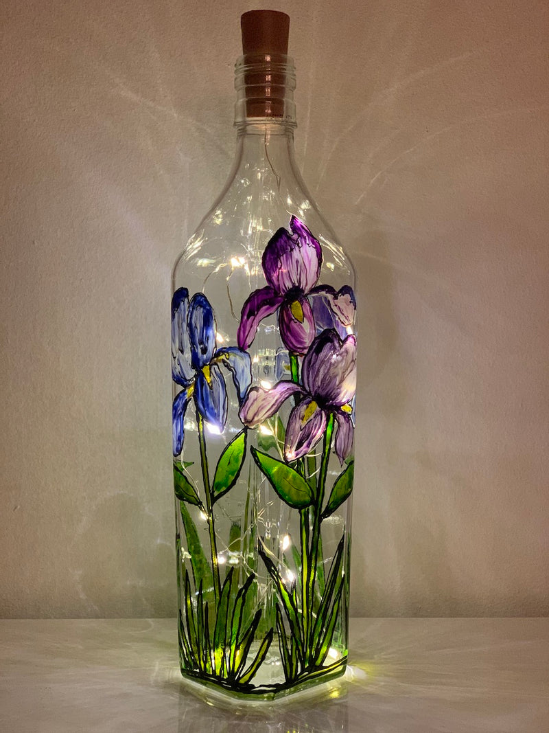Several irises stand in a garden. Large blue and purple petals show lots of texture. Painted on a recycled bottle with acrylic paint. The bottle is lit with fairy lights from within.
