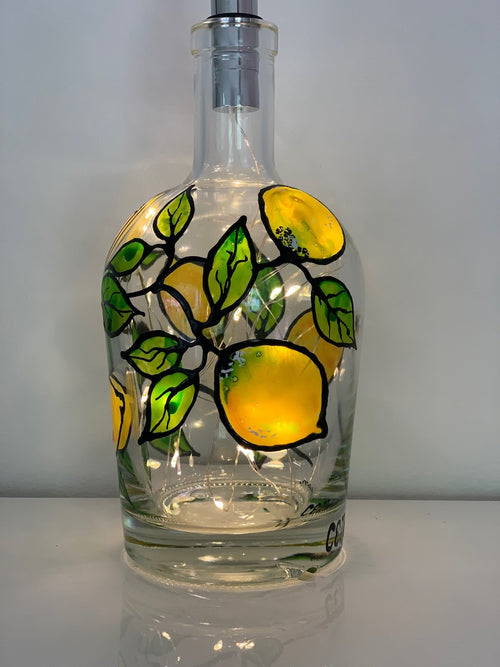 Shiny yellow lemons adorn a green branch that wraps around the bottle. Painted on a recycled bottle with acrylic paint. The bottle is lit with fairy lights from within.