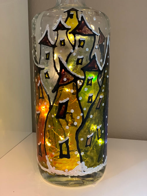 A village of colourful wavy shaped homes is warmly light at night while snow falls, creating a layer over everything. Painted on a recycled bottle with acrylic paint. The bottle is lit with fairy lights from within.