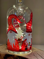 Several swedish gnomes with red suits and white beards look out from the bottles, with their eyes hidden under their hats. Painted on a recycled bottle with acrylic paint. The bottle is lit with fairy lights from within.