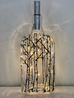 Bare white bark trees stand in a grove around the bottle. Painted on a recycled bottle with acrylic paint. The bottle is lit with fairy lights from within.