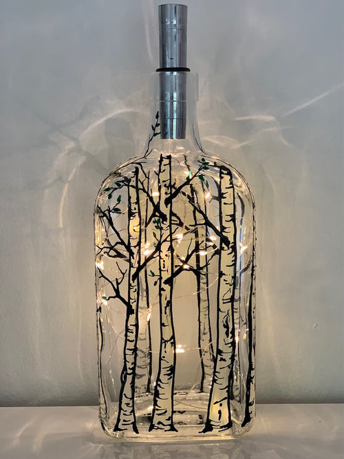 Bare white bark trees stand in a grove around the bottle. Painted on a recycled bottle with acrylic paint. The bottle is lit with fairy lights from within.