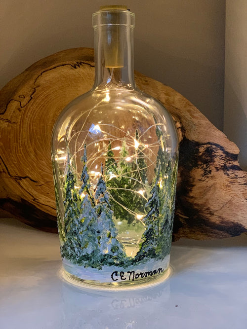 A forest of nicely textured pine trees are heavily laden with white snow.Painted on a recycled bottle with acrylic paint. The bottle is lit with fairy lights from within.