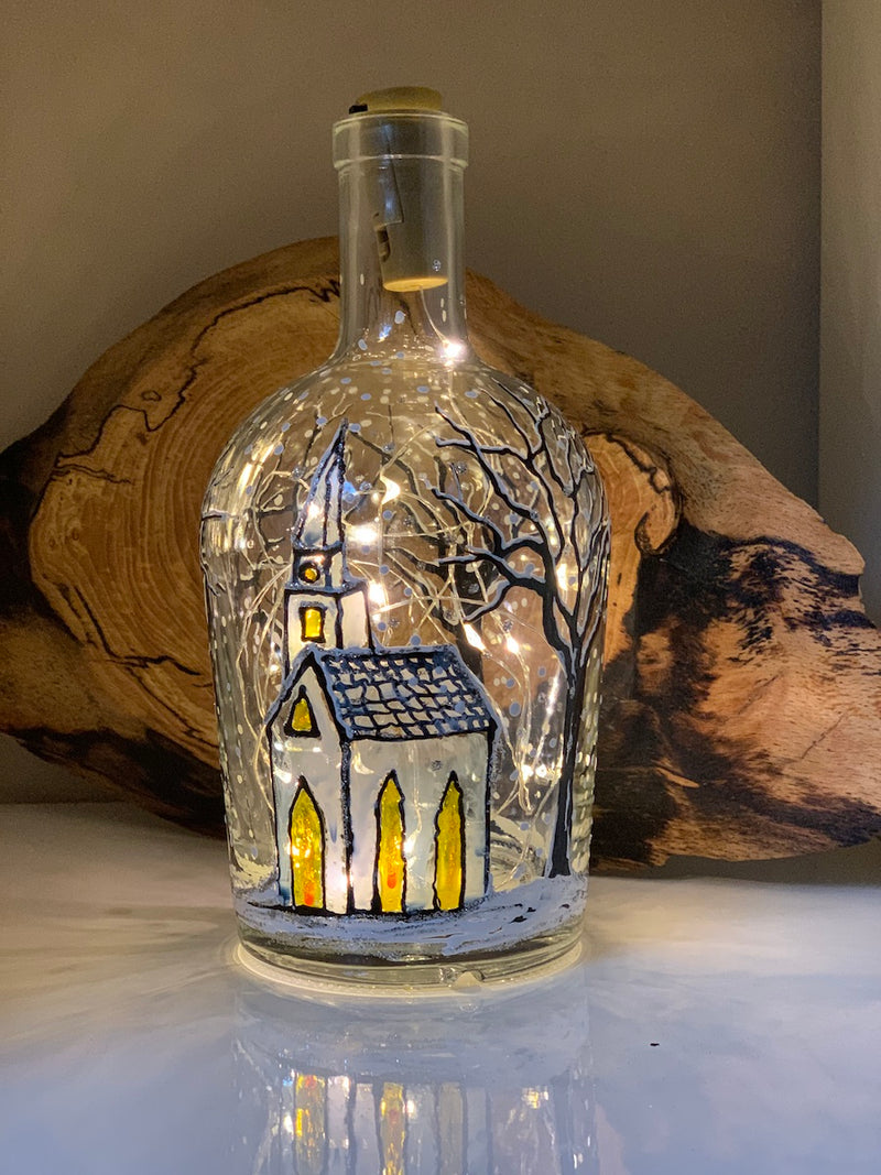 A church with a tall steeple and glowing windows stands under bare trees covered with snow.  Snowflakes fall. Painted on a recycled bottle with acrylic paint. The bottle is lit with fairy lights from within.