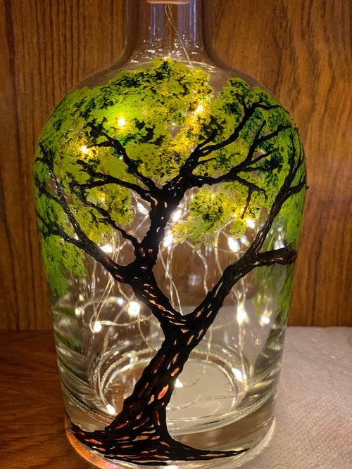 An oak tree with knotted texture and green leaves reaches from bottom to top on this bottle. Painted on a recycled bottle with acrylic paint. The bottle is lit with fairy lights from within.